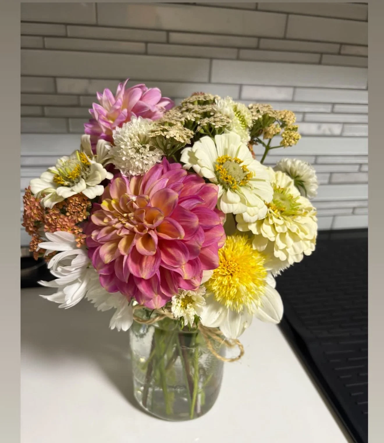 A colorful bouquet of flowers in a clear glass vase on a white surface with a brick wall background.