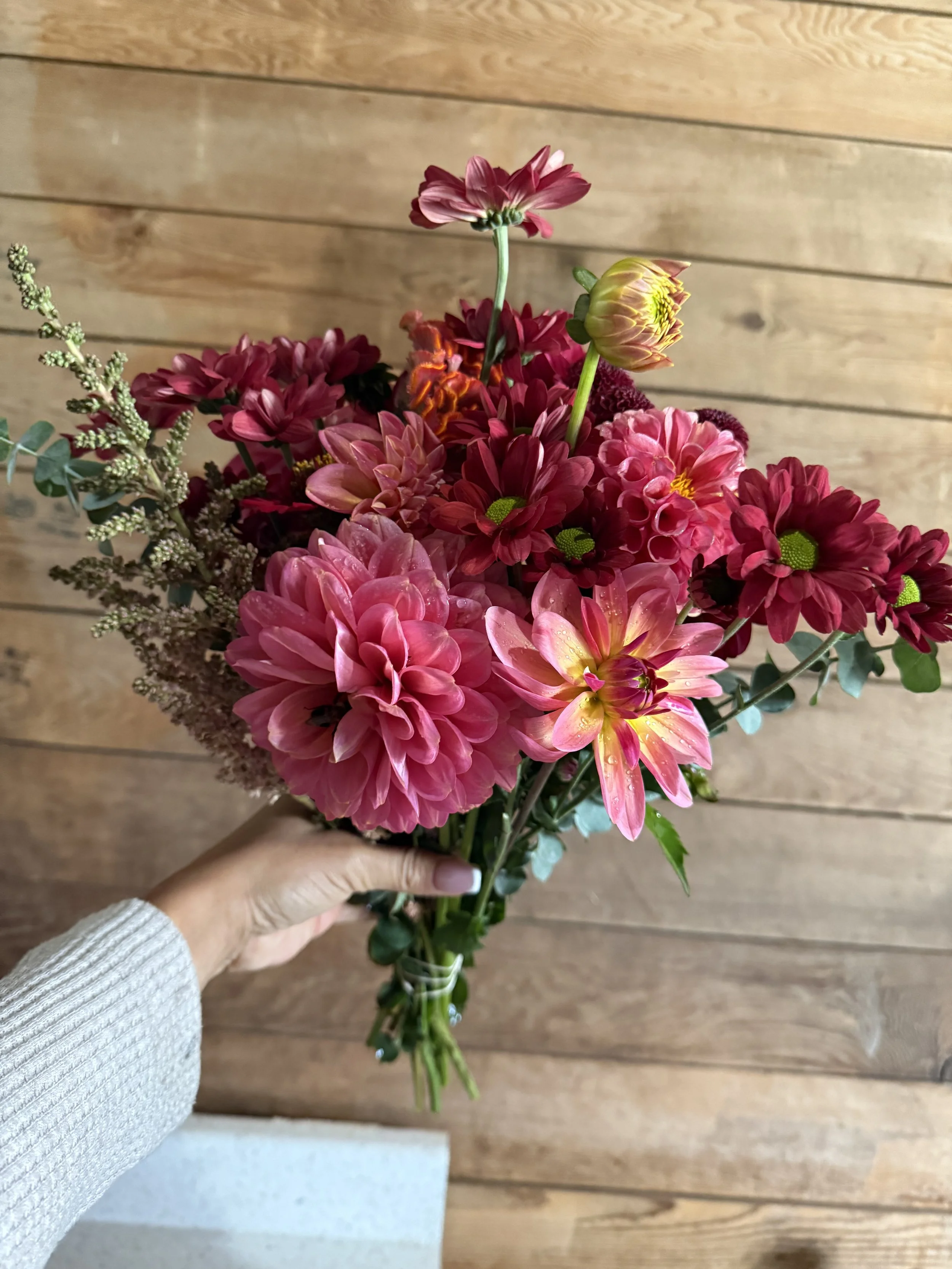 A hand holding a colorful bouquet of pink, red, and peach flowers with green foliage, against a wooden floor background.