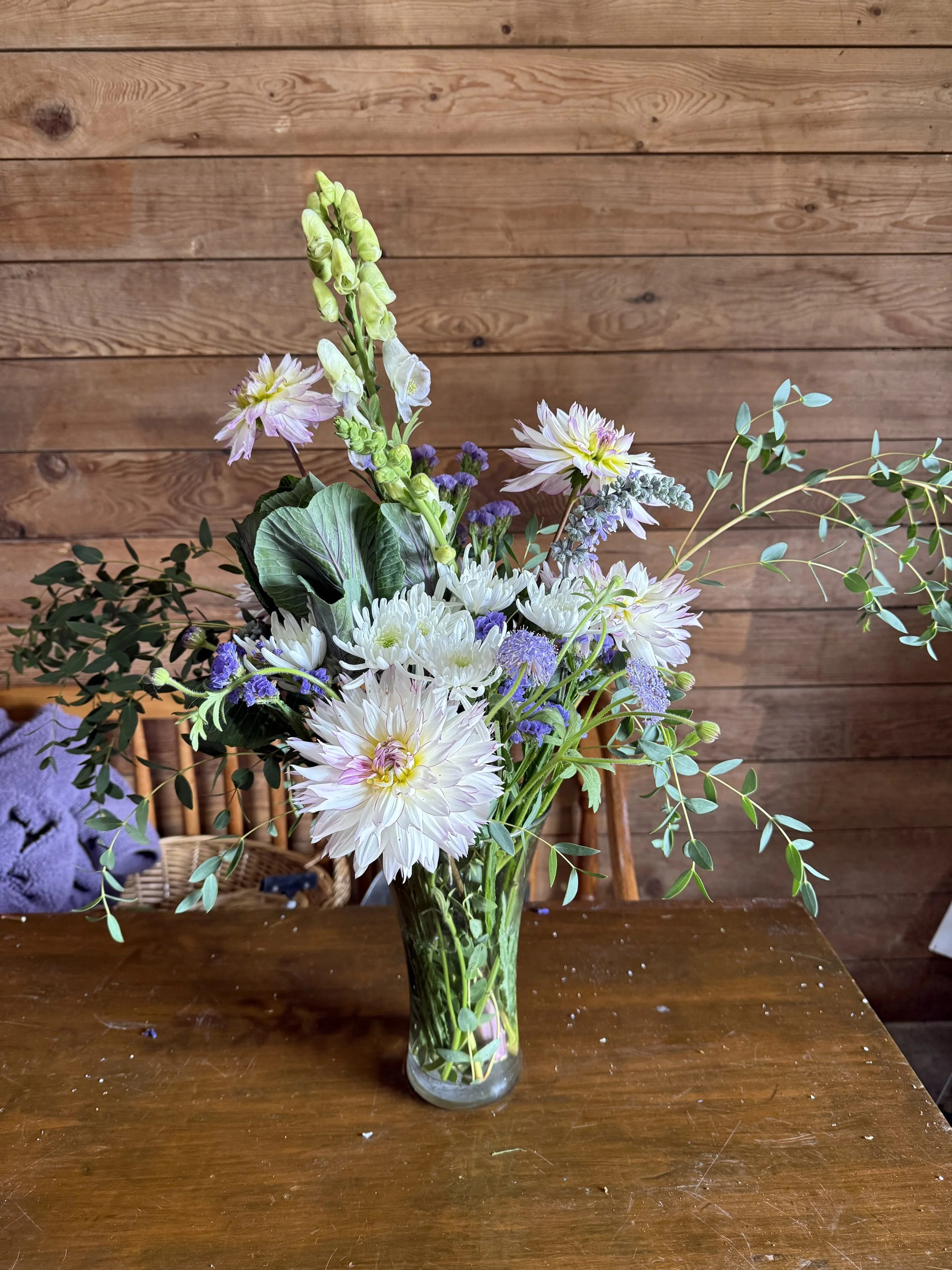A tall clear glass vase containing a mixed bouquet of white, purple, and green flowers and foliage sitting on a wooden table against a wooden wall background.