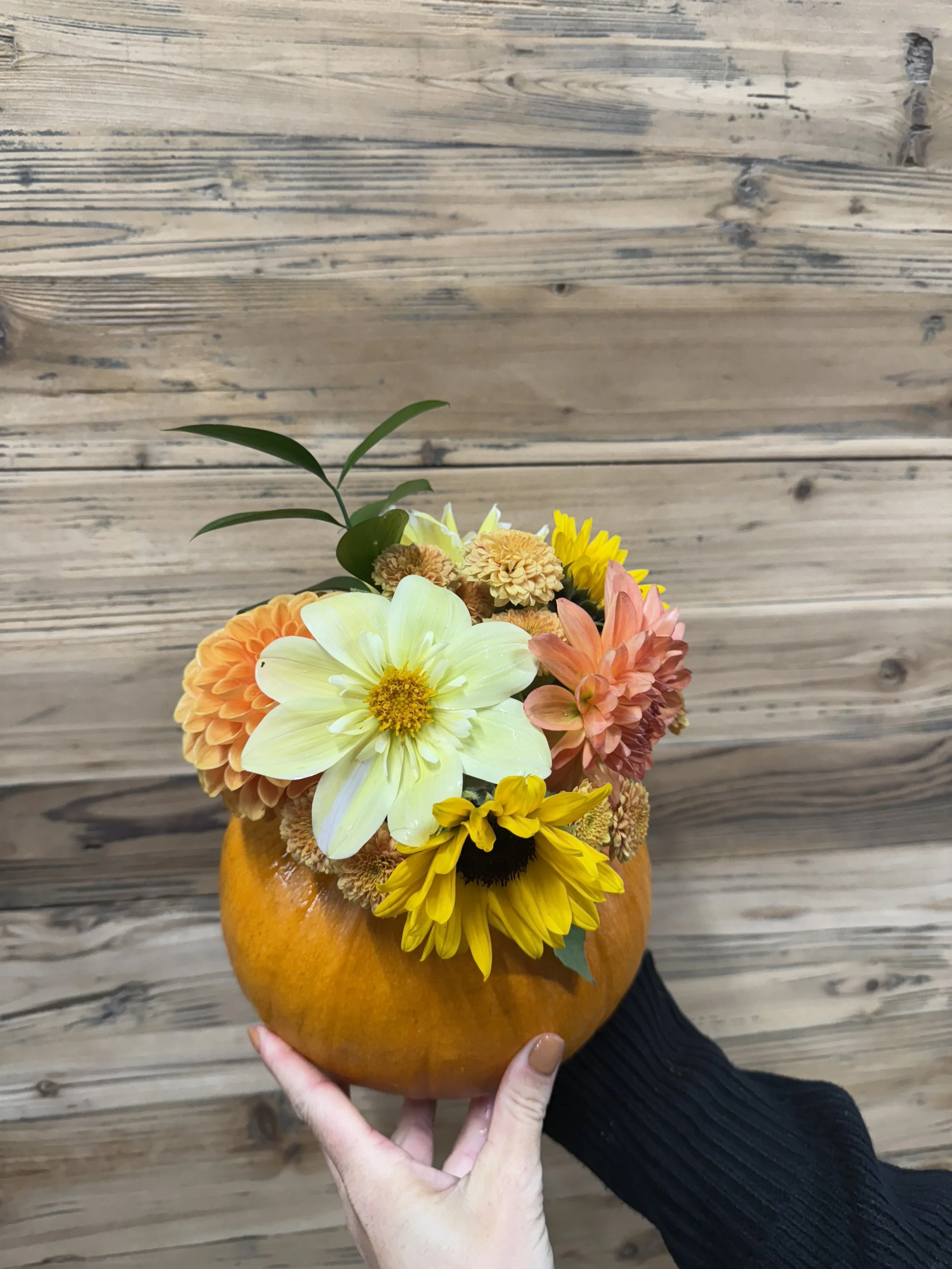 A hand holding a pumpkin filled with a colorful bouquet of flowers, including lilies, sunflowers, dahlias, and other blooms, against a wooden background.