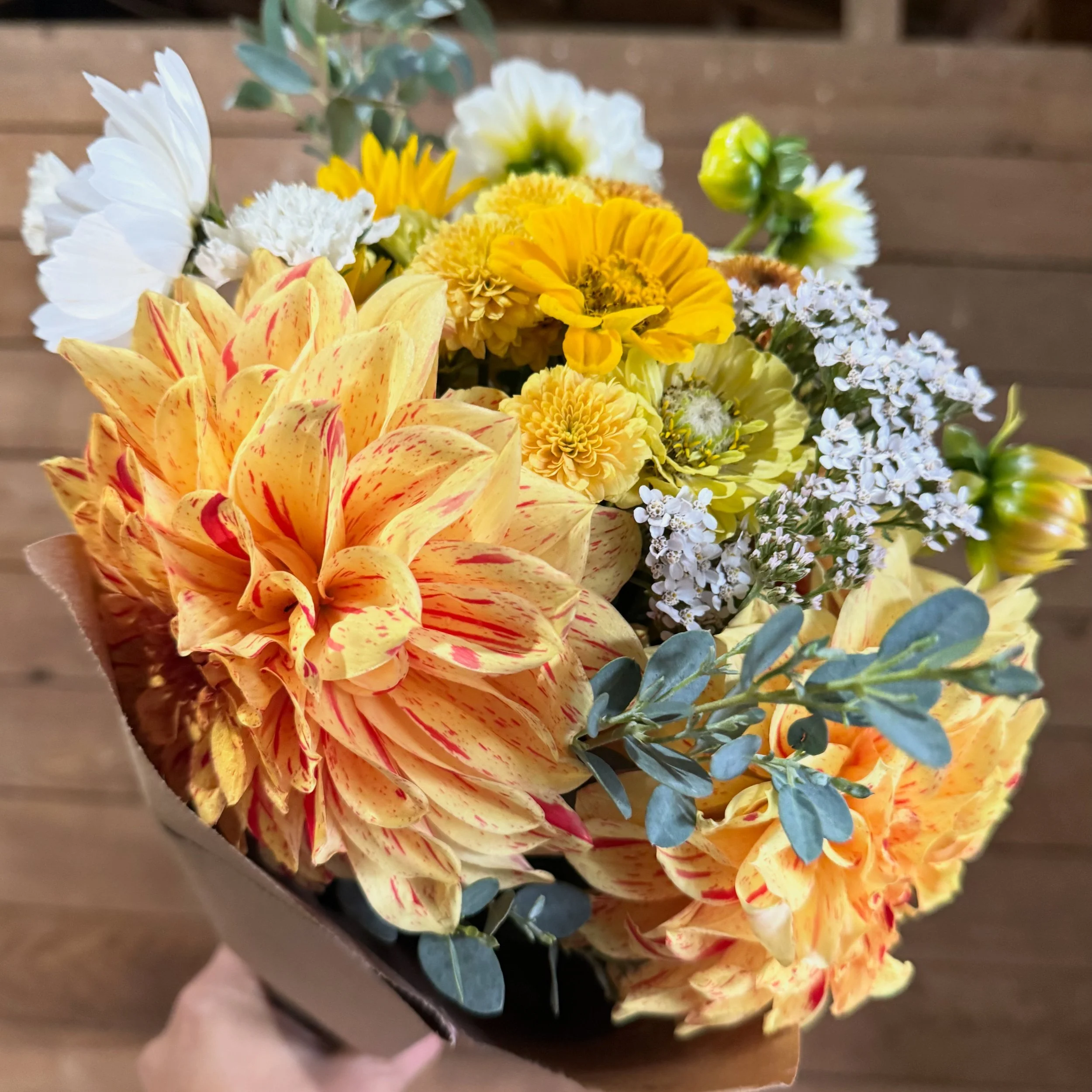 A bouquet of yellow and white flowers with large orange and red streaked dahlias, white and yellow small filler flowers, and eucalyptus leaves in brown wrapping.