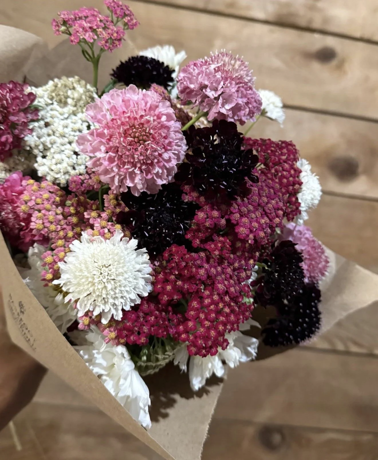 A bouquet of pink, white, and dark purple flowers wrapped in brown paper, set against a wooden background.