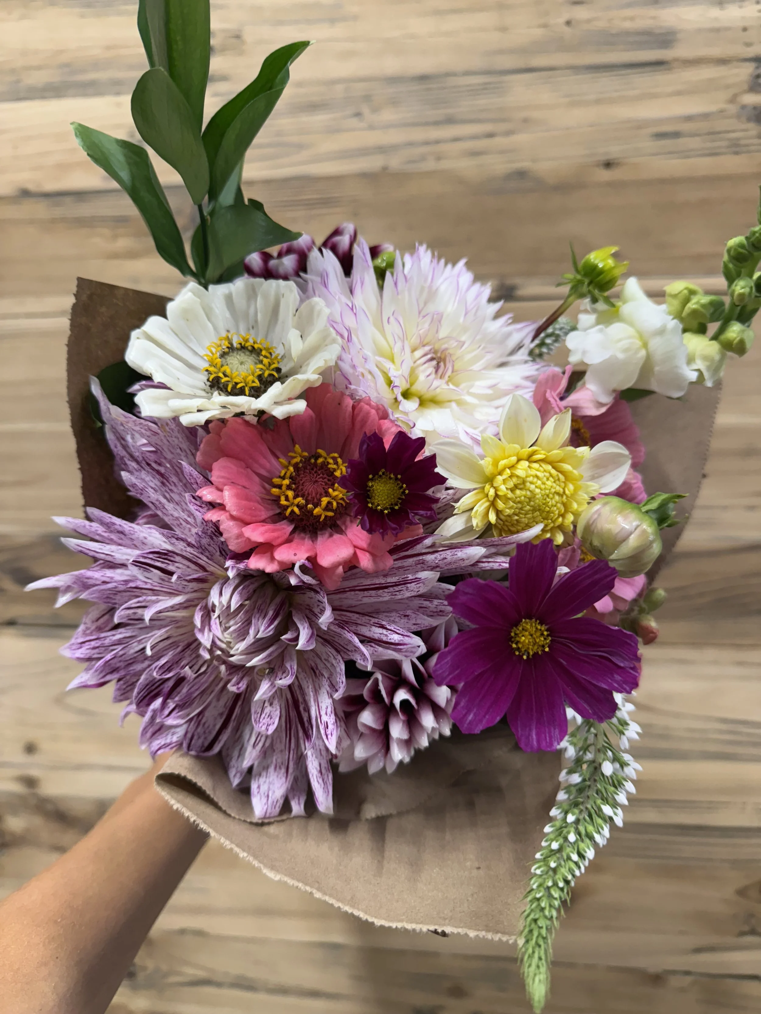 A colorful bouquet of various fresh flowers wrapped in brown paper, held over a wooden floor background.