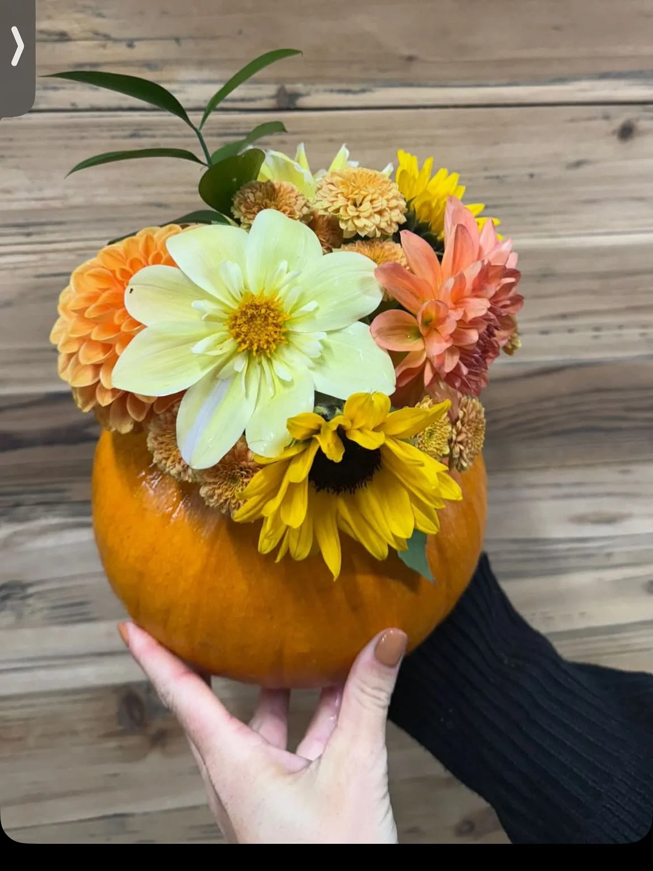 A pumpkin-shaped vase with colorful flowers including sunflowers, daisies, and dahlias, being held by a person with painted nails over a wooden background.