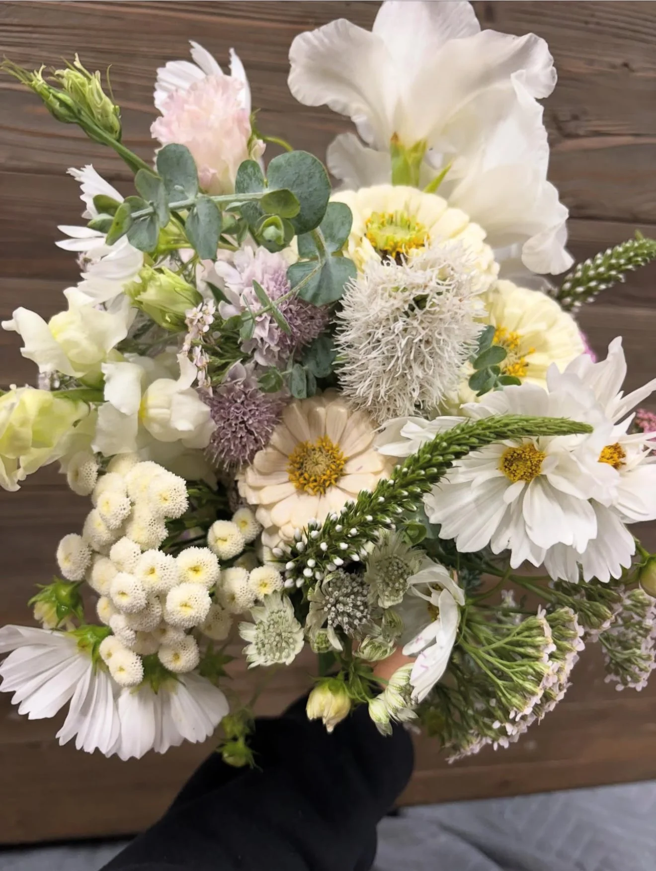 A bouquet of white, cream, and light purple flowers with green foliage, including daisies, lavender, and other mixed blossoms, held up against a wooden background.