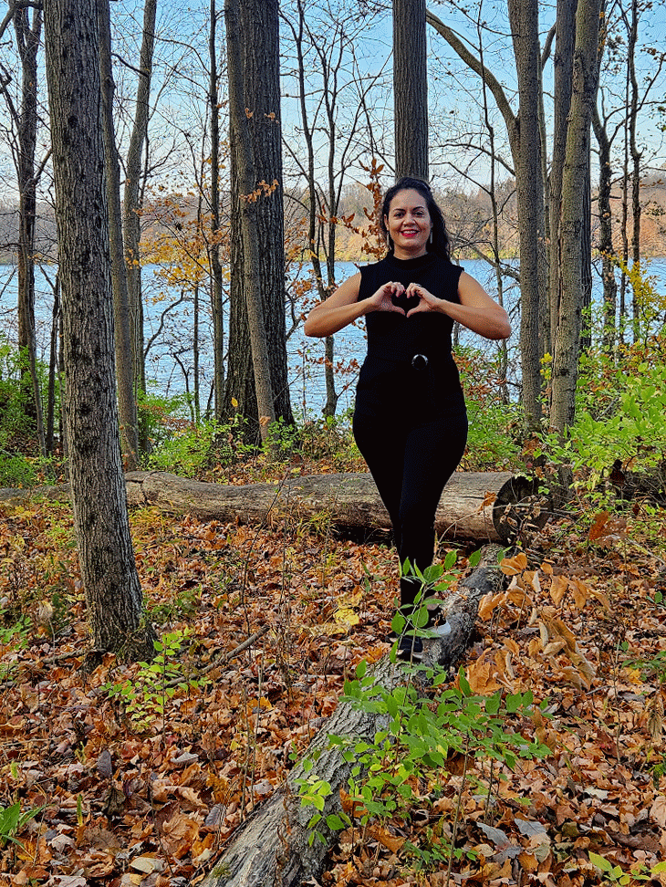 A woman standing on a fallen tree in a forest during autumn, making a heart shape with her hands and smiling.
