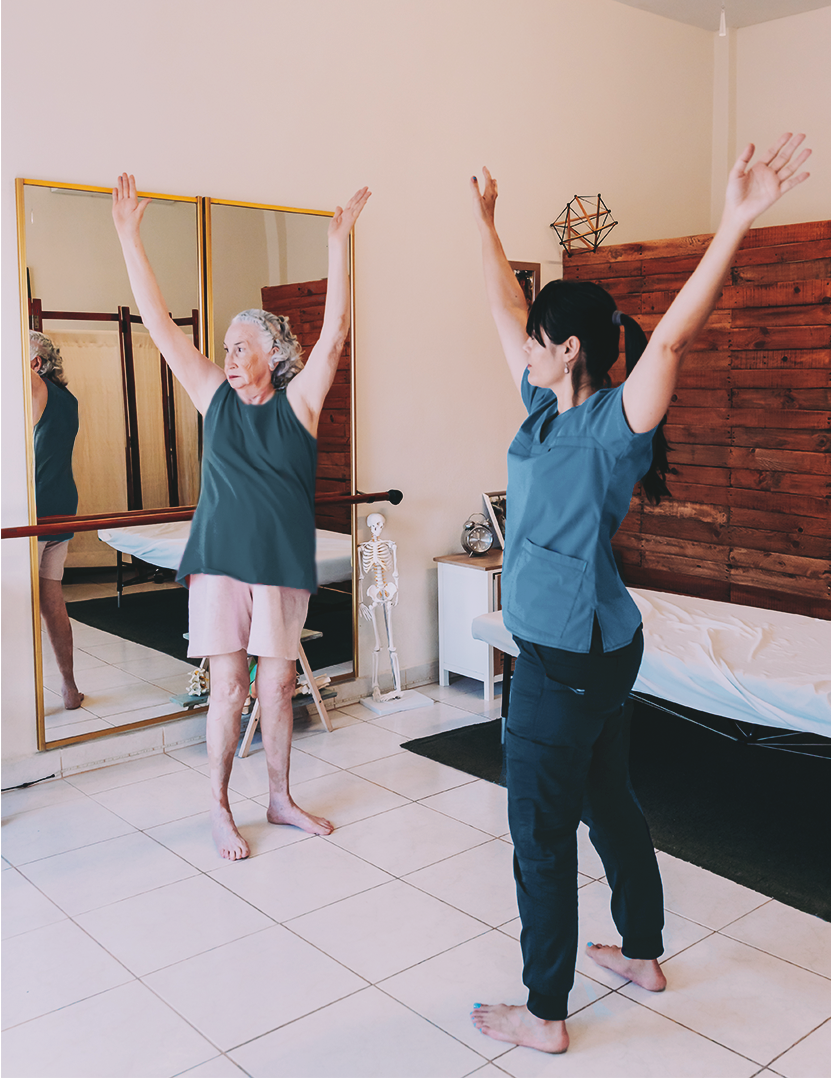 An elderly woman with gray hair practicing yoga in front of a mirror, with her arms raised, while a young woman in medical scrubs guides her.