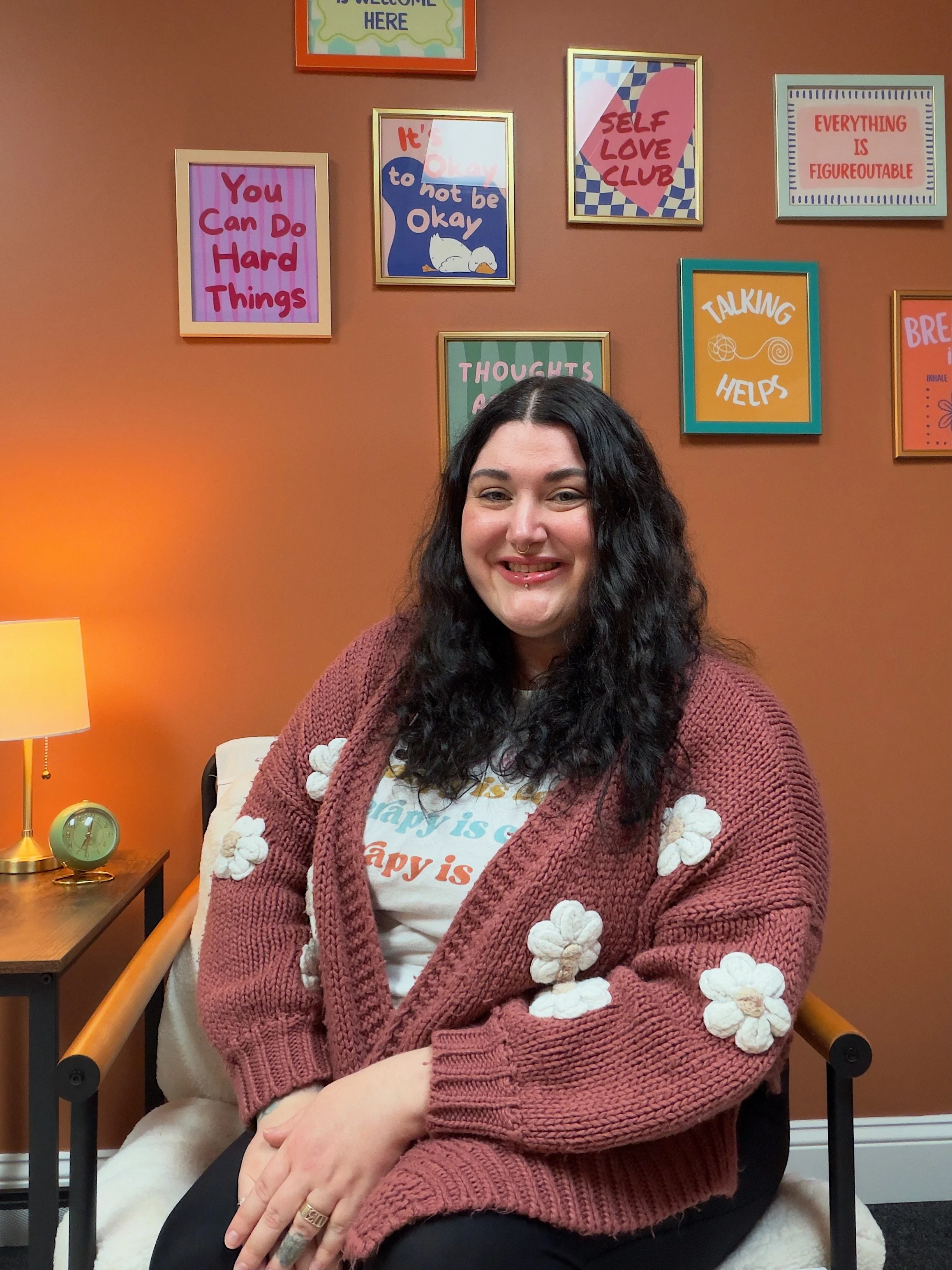 A woman with long curly black hair, wearing a maroon knitted cardigan decorated with white flowers and a white T-shirt with colorful text, is sitting in a room with an orange wall. The wall has several framed inspirational and motivational quote posters and a framed abstract art. There is a small wooden side table with a green modern clock and a gold lamp with a cream lampshade beside her.