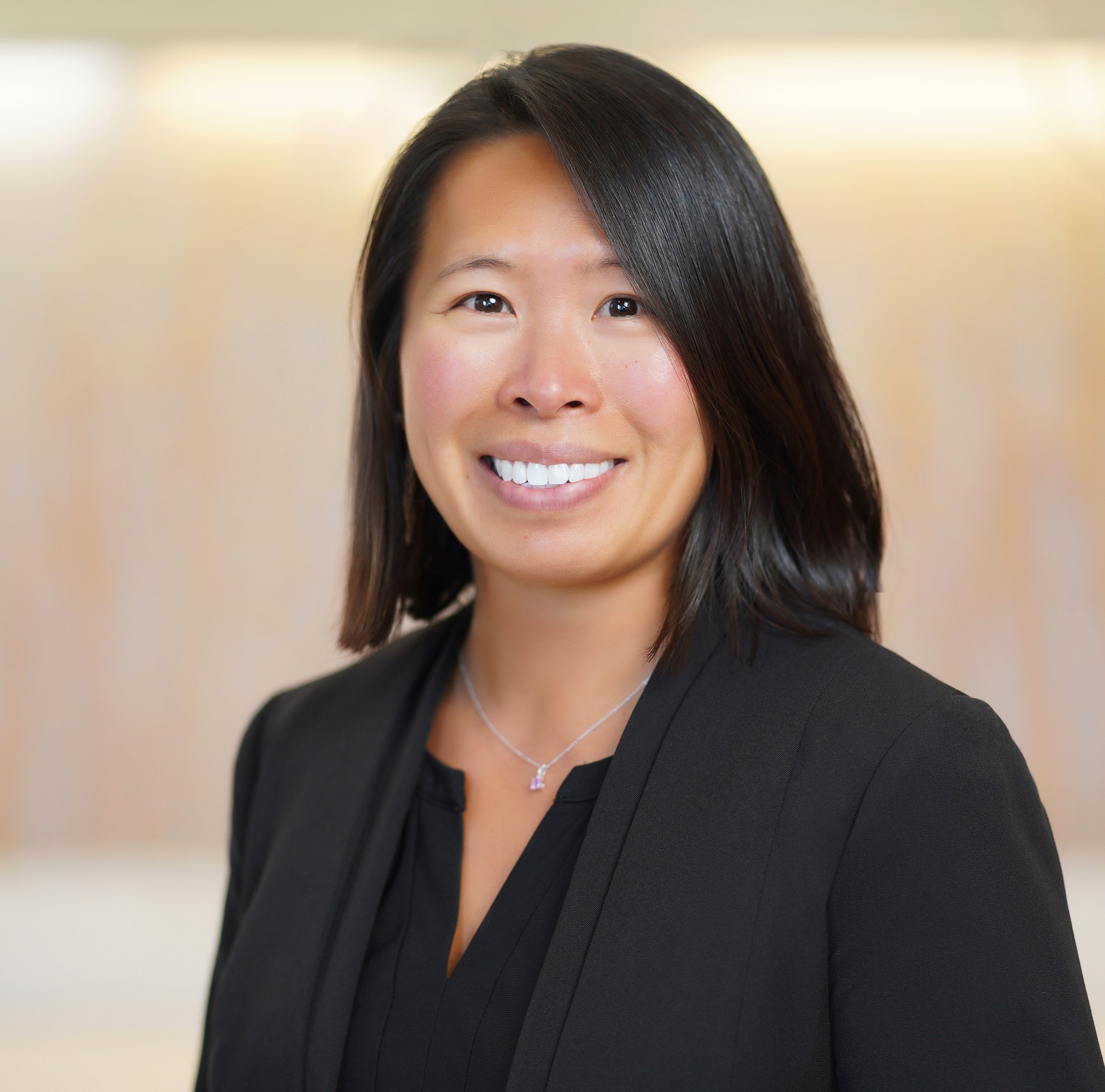 Portrait of a woman with shoulder-length dark hair, smiling, wearing a black blazer and a necklace, against a blurred beige background.