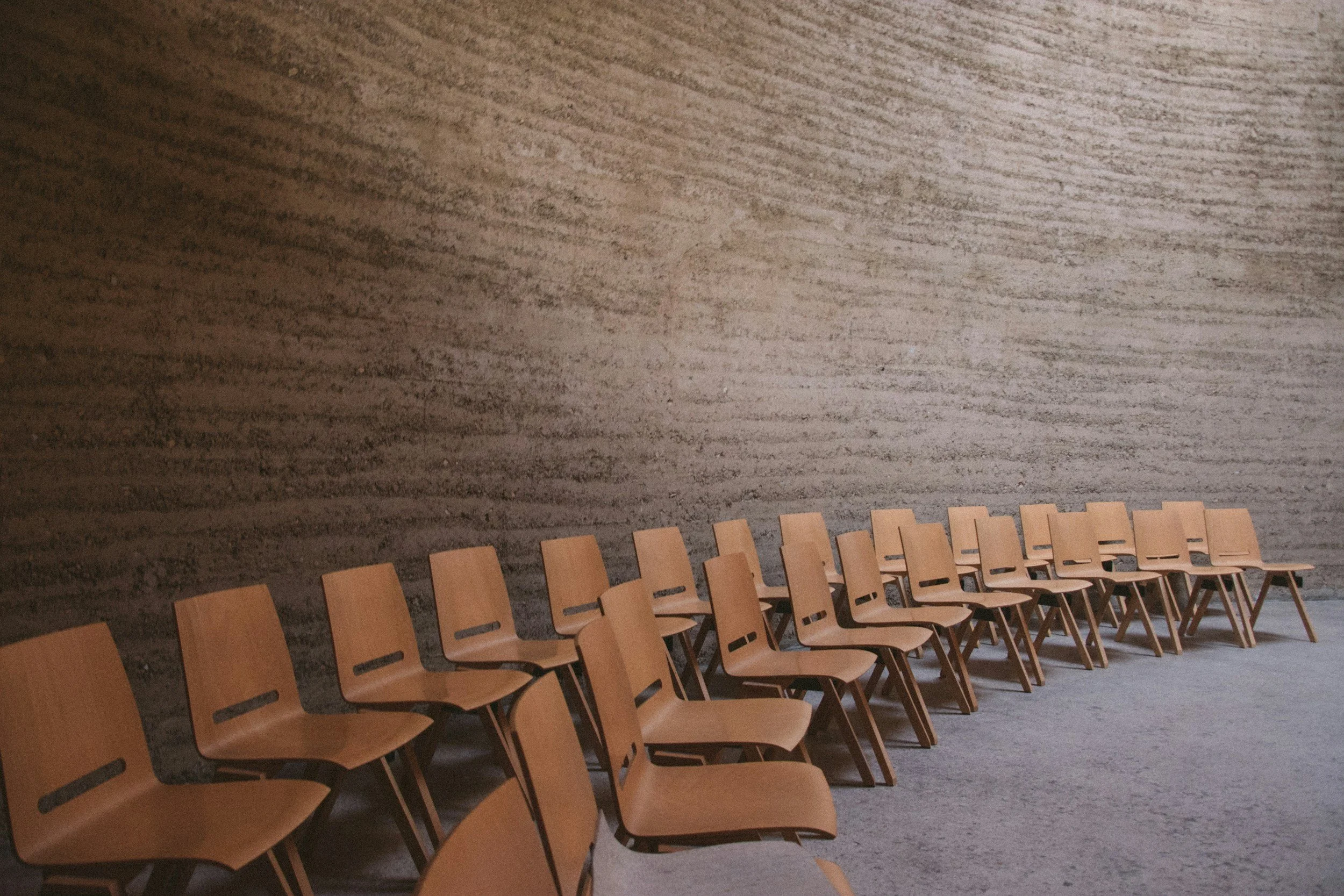 Row of wooden chairs lined along a large textured wall in an empty room.