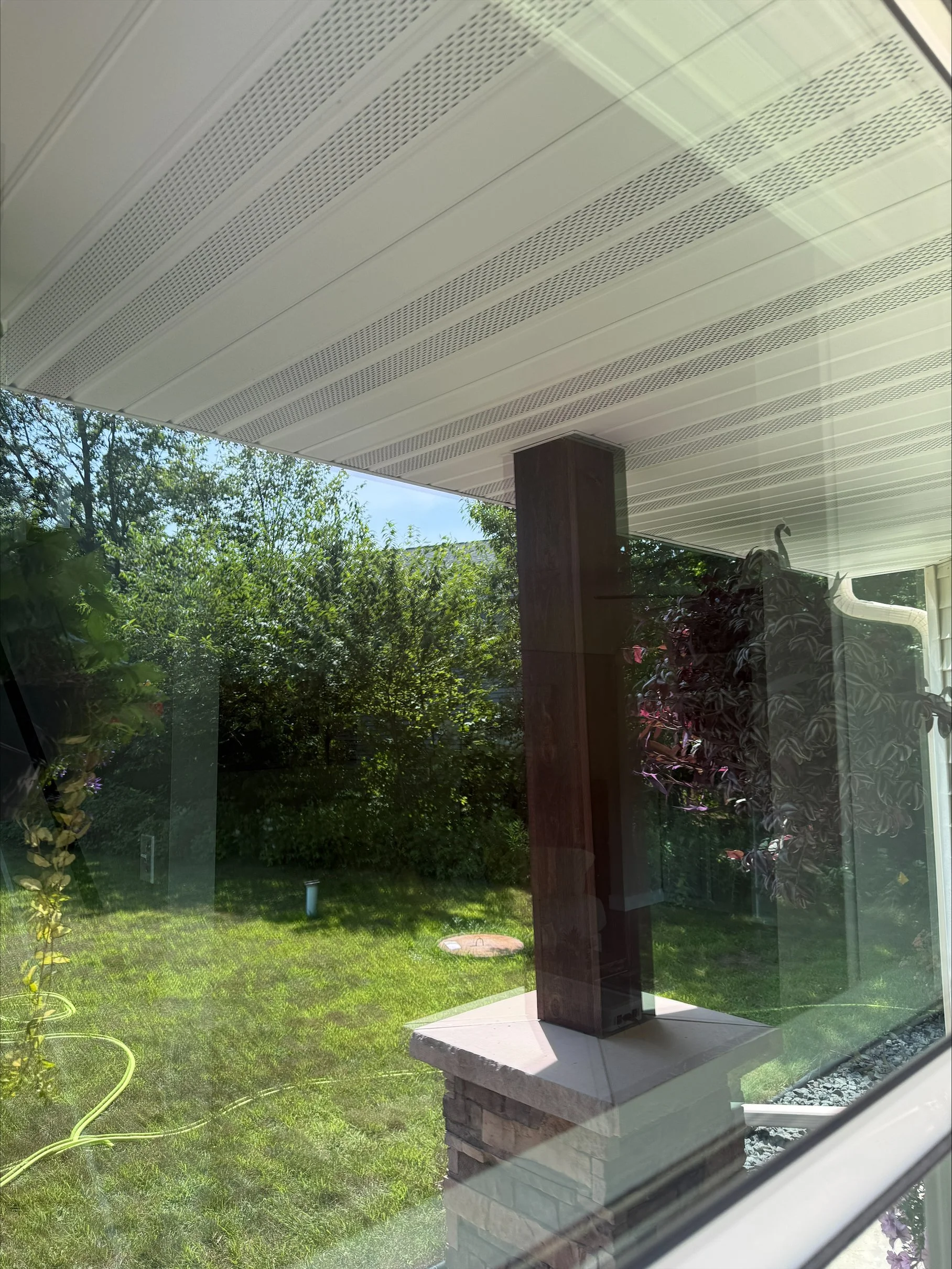 View of a backyard through a window, showing a grassy lawn, trees, and a covered porch with a wooden support post, a brick pillar, and hanging plants.