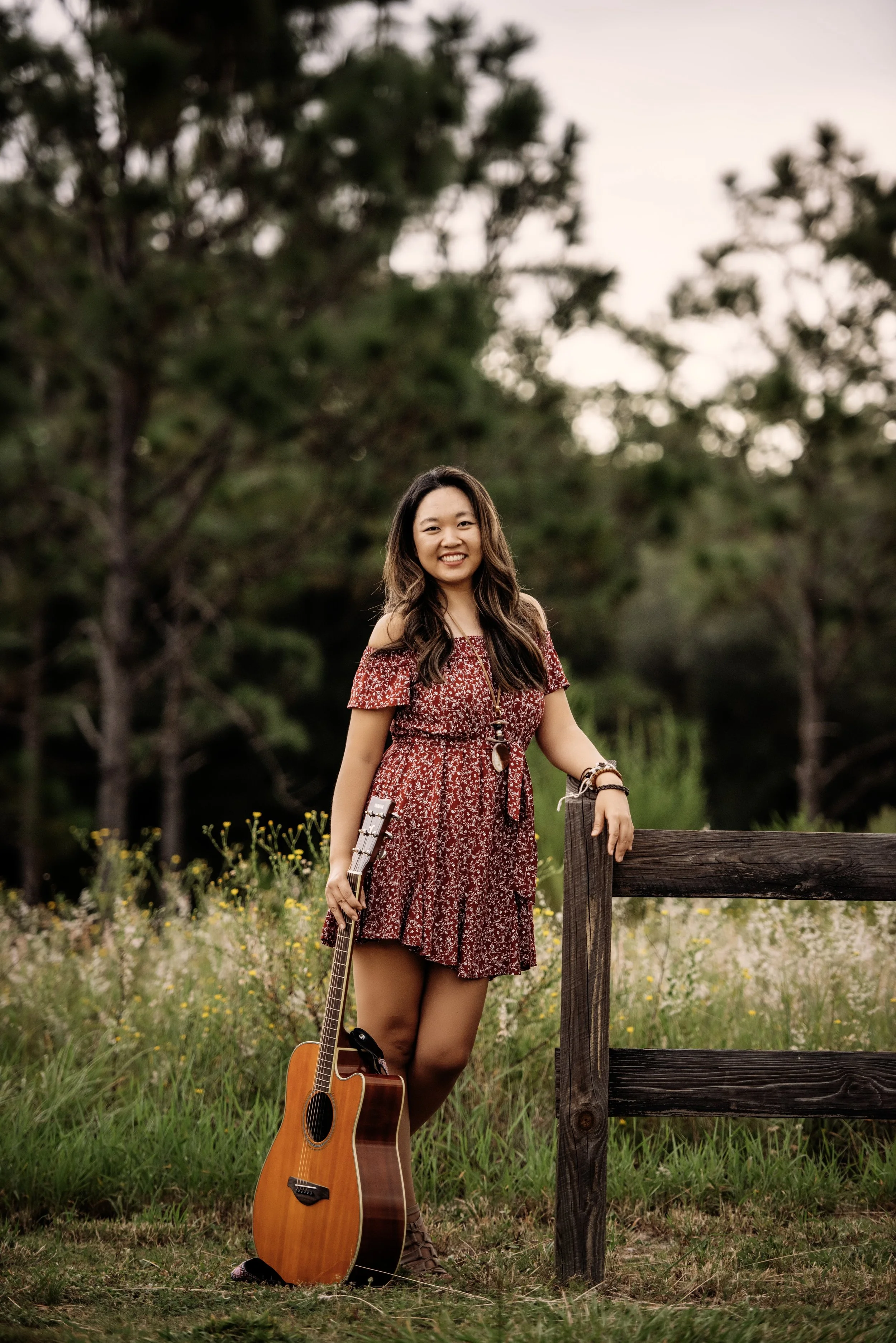 A woman in a red floral dress stands outdoors in a meadow with trees in the background, holding an acoustic guitar.