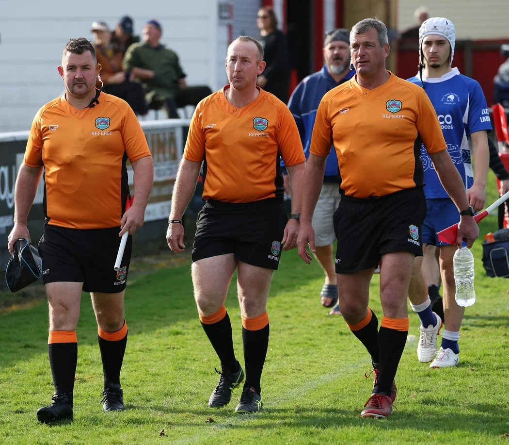Three male referees walking on a grass field, wearing orange shirts, black shorts, and black socks with orange accents, with one holding a water bottle and another carrying a small bag. They are leading a group of people, possibly during a sports eve
