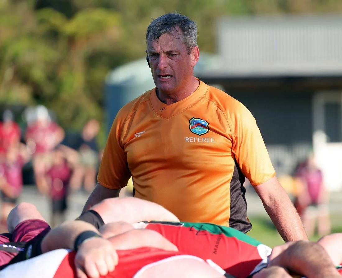 Referee in orange jersey officiating a rugby match. The referee is focused, with players lying on the ground in front of him. Blurred background with spectators and part of a building.