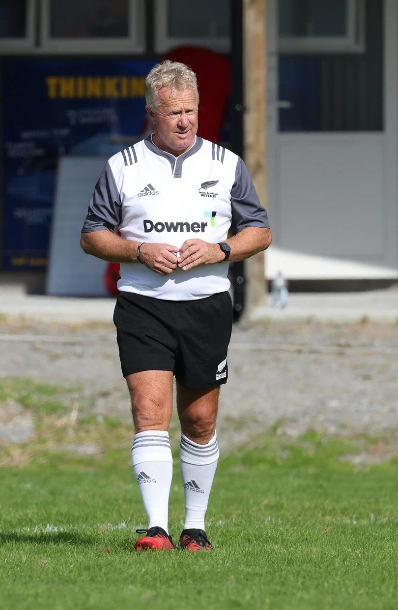 A man in a rugby referee uniform standing on grass field, with a building in the background.
