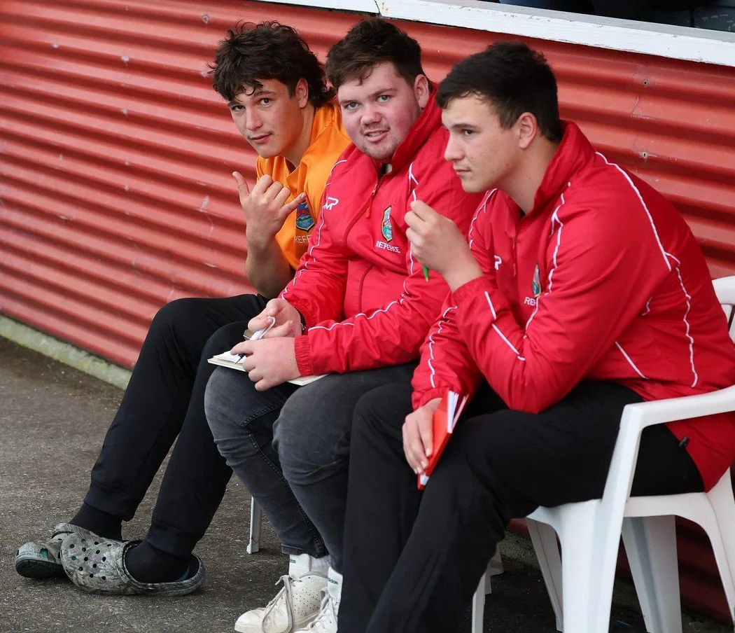 Three young men sitting on a bench outside, wearing red jackets and black pants, with one in an orange shirt, against a red corrugated metal wall.