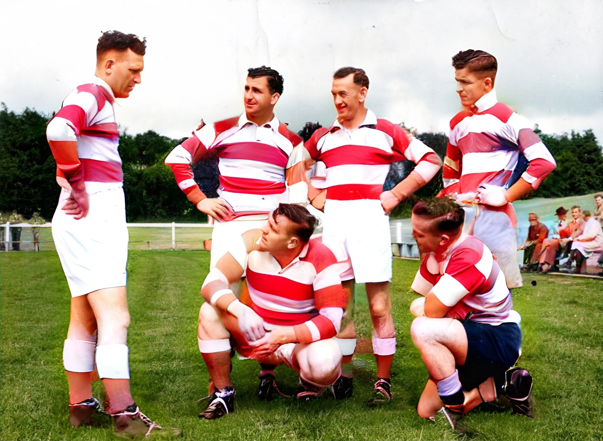 A group of seven rugby players in red and white striped jerseys and shorts on a grassy field, with some players standing with hands on hips and two crouching or kneeling, engaged in a huddle or team discussion during a match or practice, with spectat