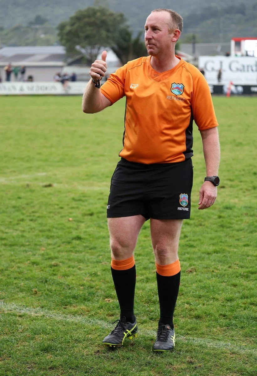 A male soccer referee standing on a grass field, wearing an orange referee jersey, black shorts, and black knee-high socks with orange accents, giving a thumbs-up gesture.