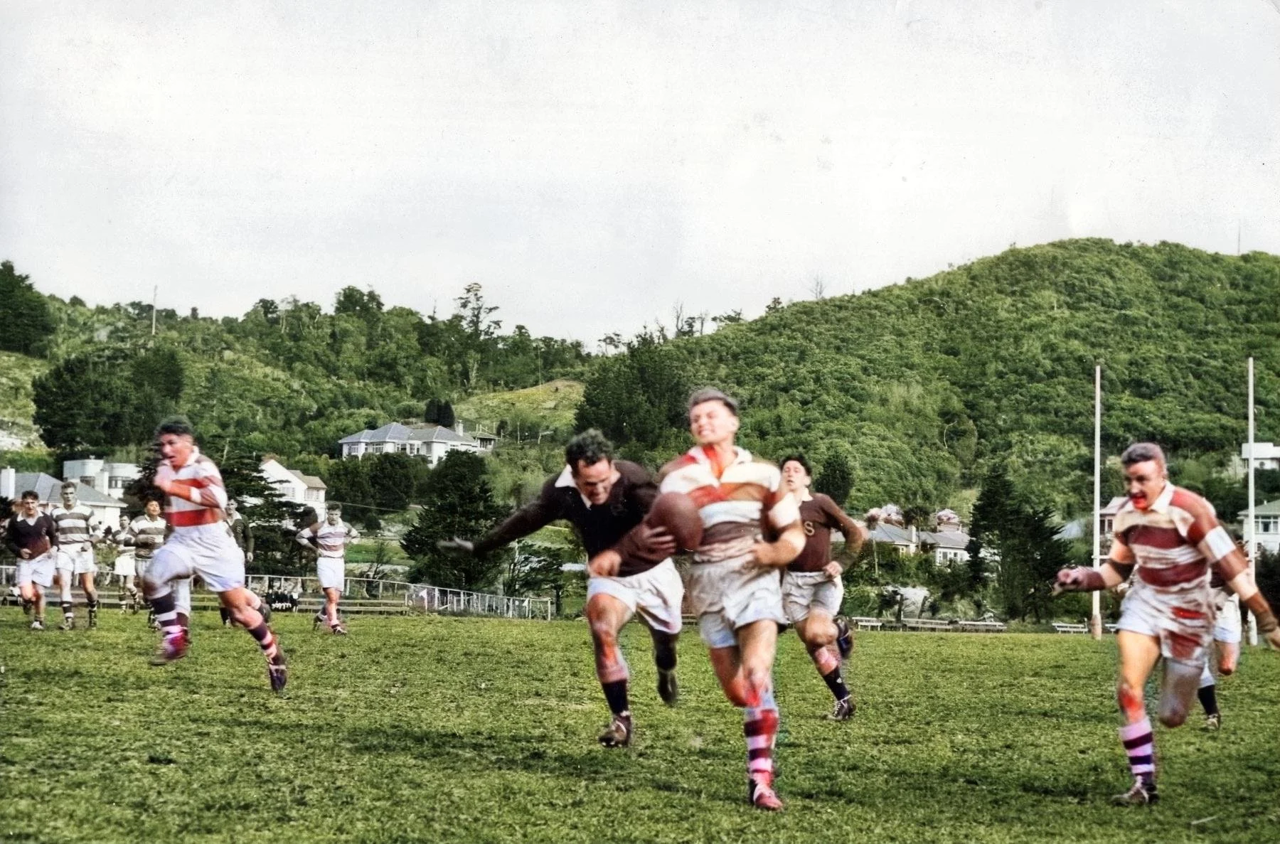 A rugby game with players running on a grass field, some with bloodstains, a player holding a rugby ball. Hills and houses are in the background.