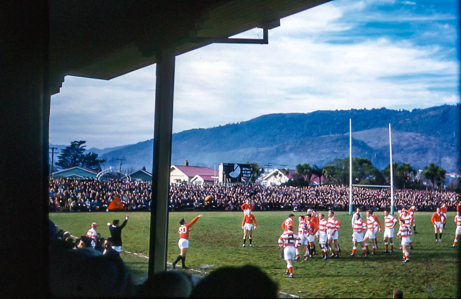 A rugby game taking place on a grassy field with players in red and white striped uniforms. A crowd of spectators is seated in the background under a partly cloudy sky, with mountains visible in the distance.