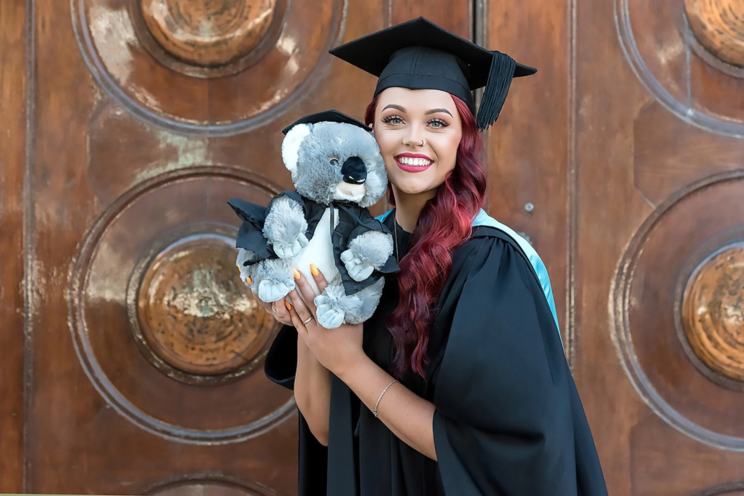 A young woman in a black graduation gown and cap holding a gray teddy bear in front of a stained glass window.
