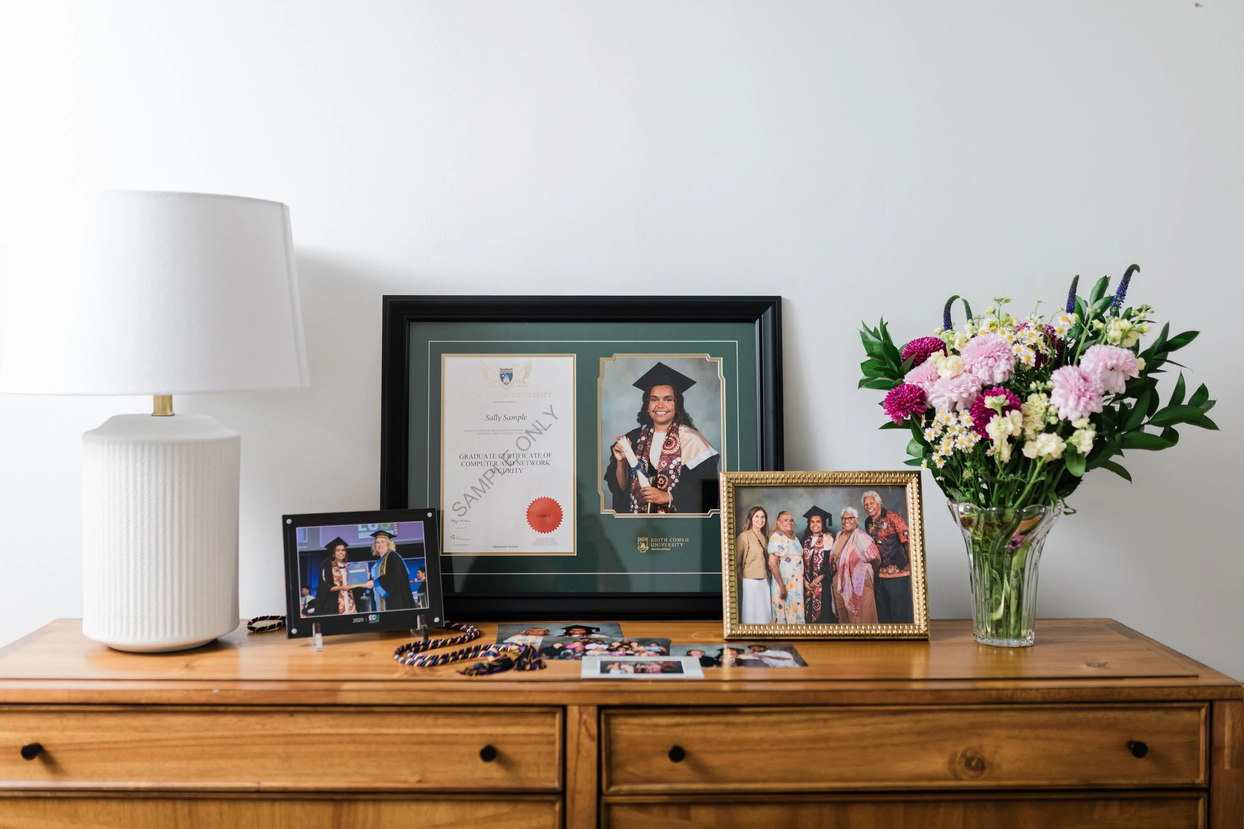 A wooden dresser with a white lampshade, graduation photos including a framed diploma, a portrait of a young woman in cap and gown, a group photo, a digital photo frame, a bunch of colorful flowers in a glass vase, and some graduation tassels.