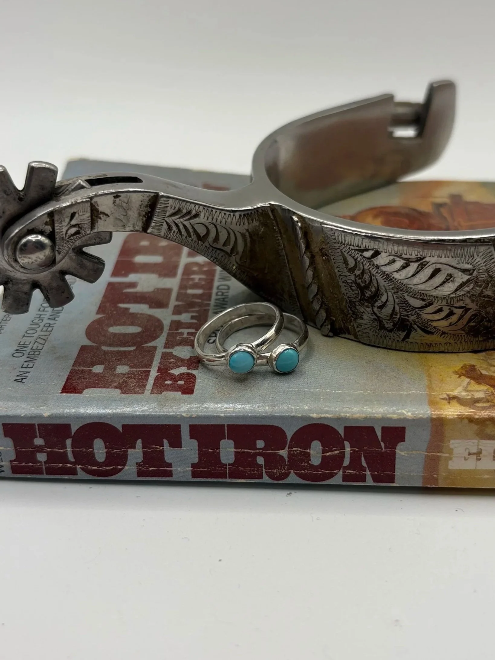 A pair of silver rings with turquoise stones, a silver bracelet with engraved designs, and a vintage book titled 'Hot Iron' on a white surface.