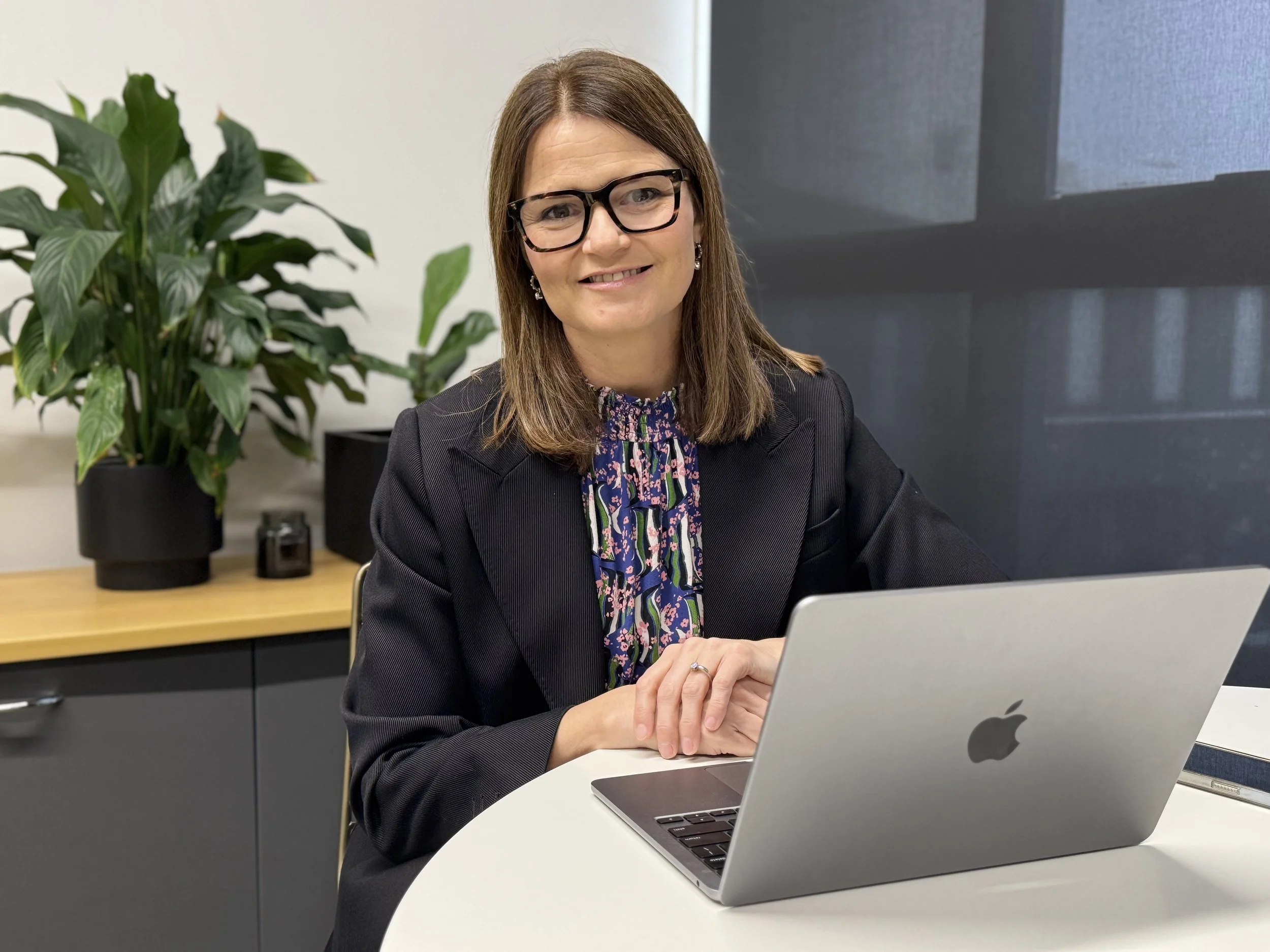 A woman with shoulder-length brown hair, wearing glasses and a black blazer, sitting at a white table with a silver MacBook laptop in an office setting, with a large green plant and a window with blinds in the background.