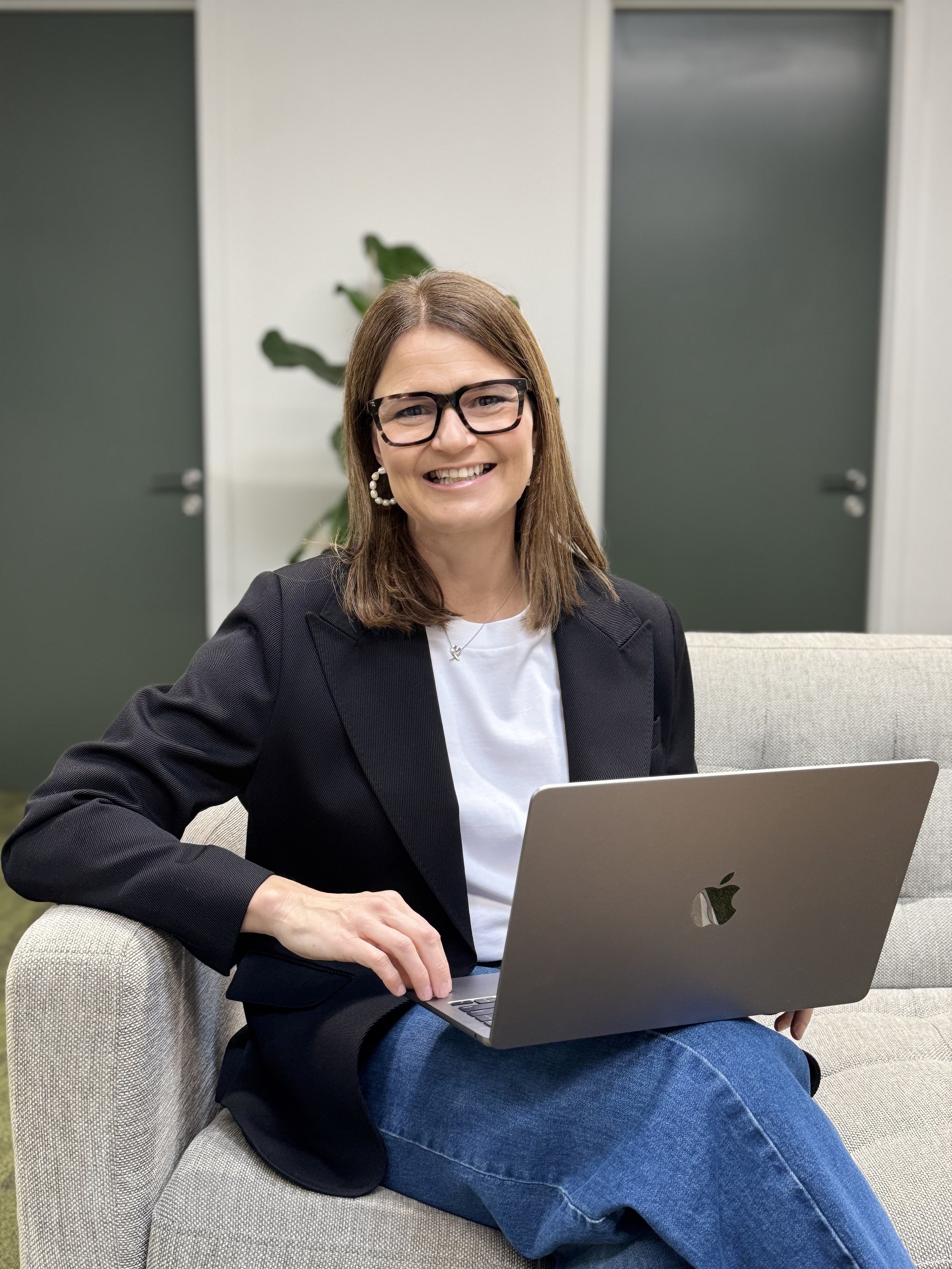 A woman with brown hair, glasses, and pearl earrings sitting on a beige couch, smiling while using a silver MacBook laptop in an office setting.