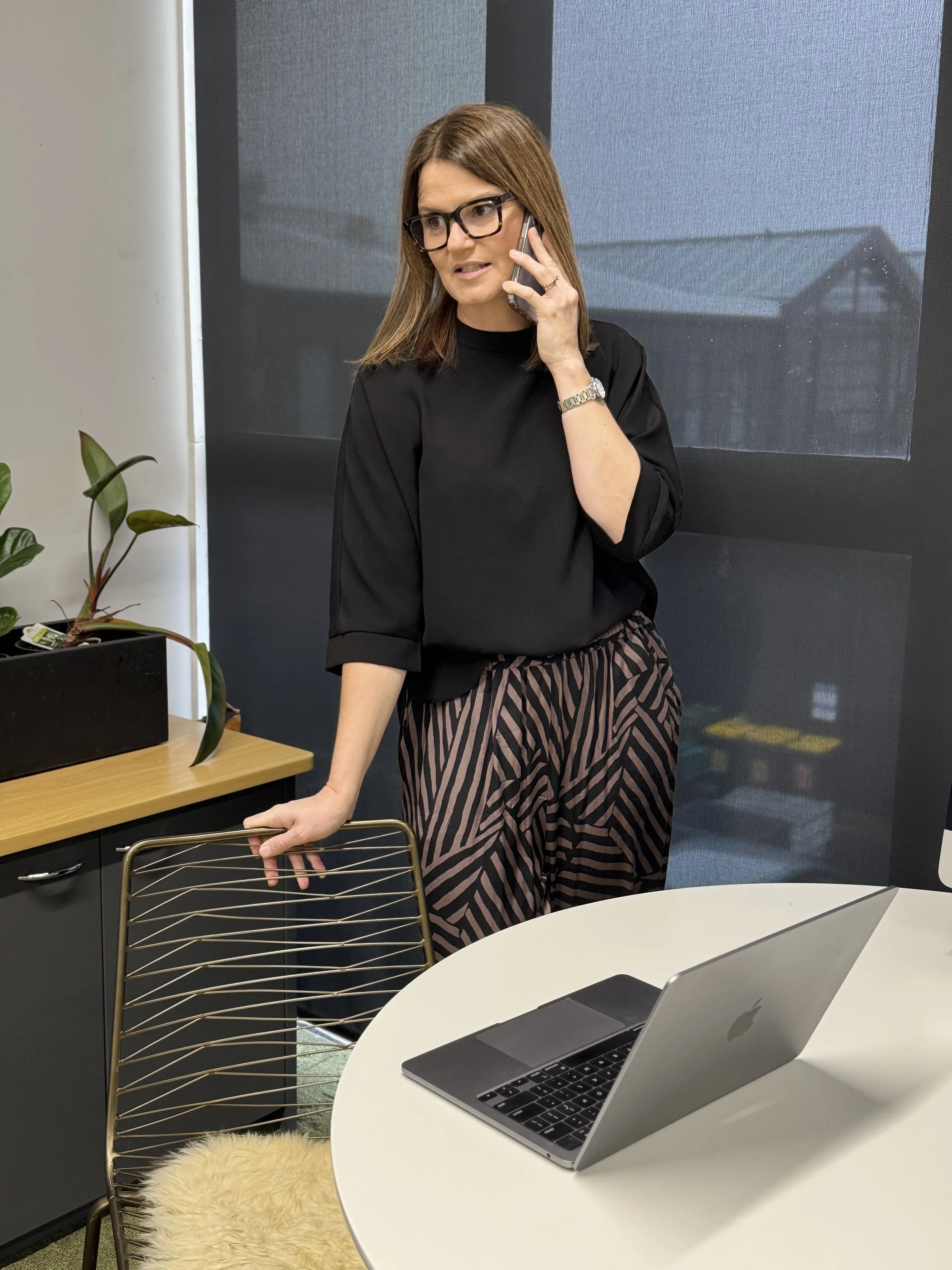 A woman with brown hair, glasses, and a black top is talking on a cell phone in an office setting, standing beside a white round table with a silver open laptop. She is holding a chair with one hand and has a plant and a cabinet behind her.