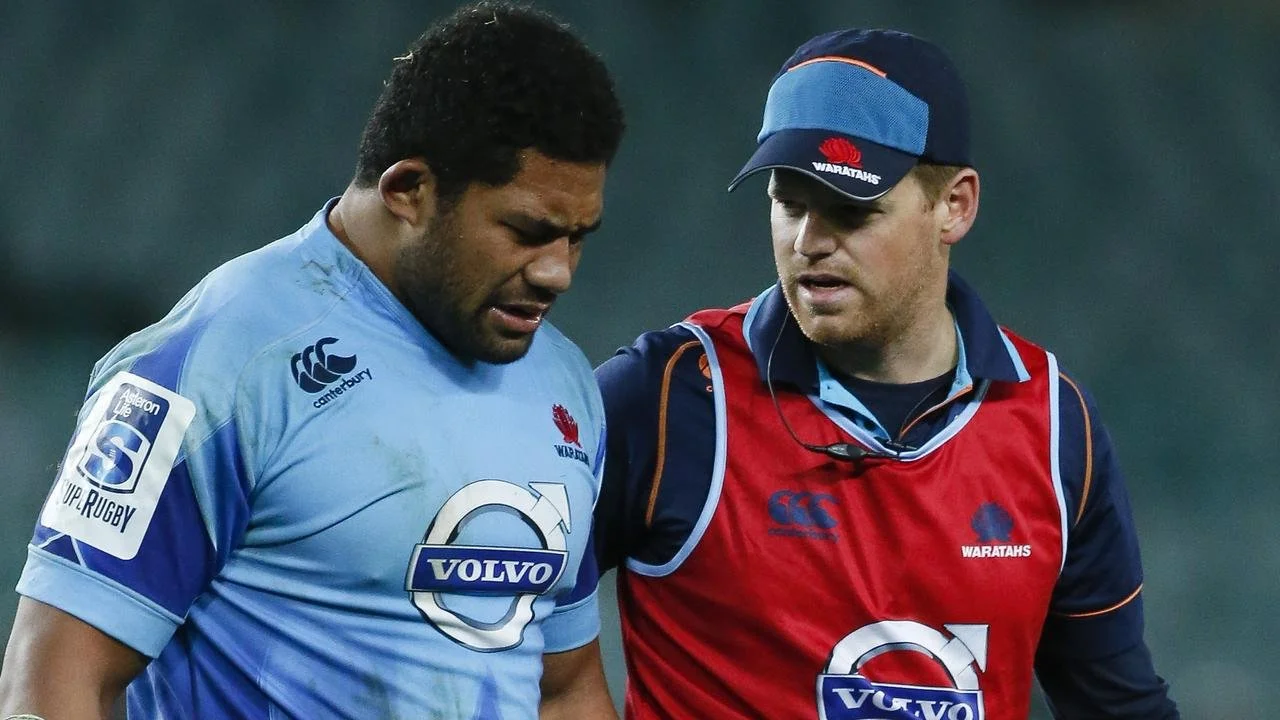 Two rugby players, one in a light blue jersey and the other in a red and dark blue jersey, appear to be in discussion on the field. The player in blue has short curly hair and the player in red wears a cap. Both jerseys have the Volvo logo and the Canterbury brand, and the red jersey also features a Waratahs logo.