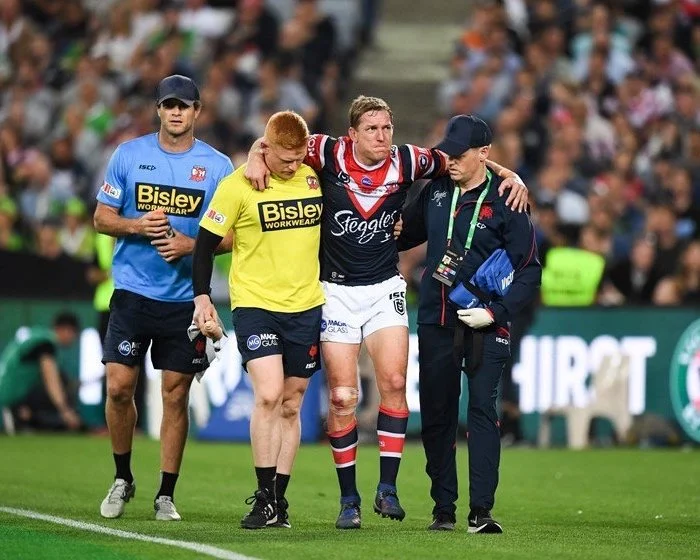 A rugby player with a bandaged leg being helped off the field by medical staff, accompanied by supporters and team personnel.