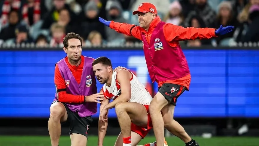A sports injury scene at a football game with medical staff assisting an injured player on the field while a coach or referee gestures with arms outstretched.