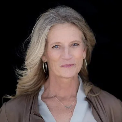 A portrait of an older woman with shoulder-length gray hair, wearing a light blue blouse and a brown jacket, against a black background.