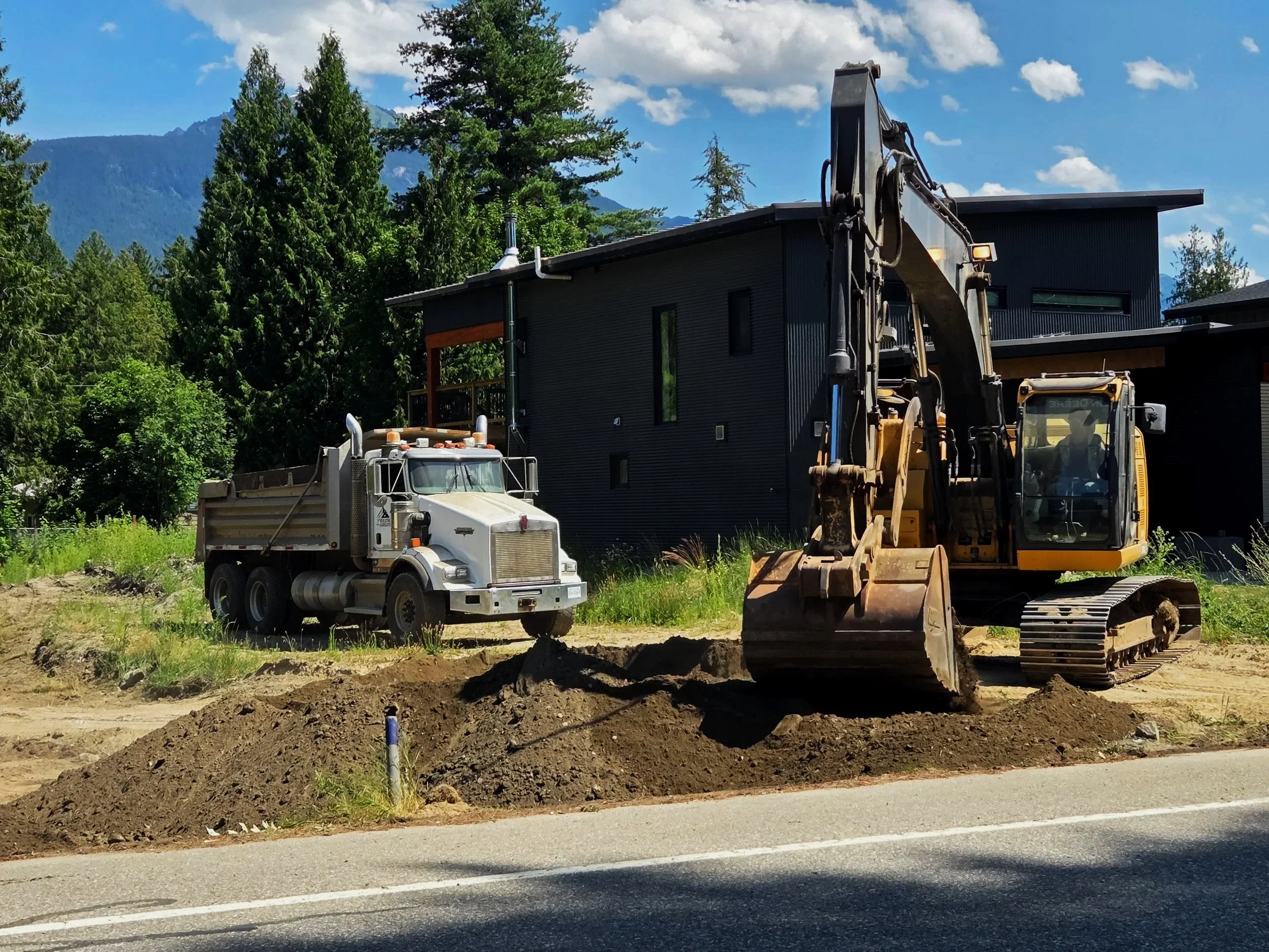Excavator moving dirt to prepare a construction lot in Revelstoke, BC, with a white Kenworth dump truck parked in the background.