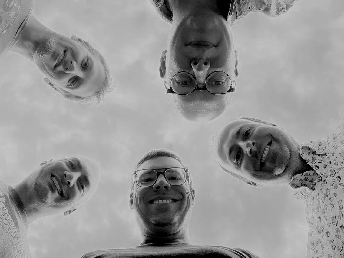 A group of five people looking down into the camera, which is positioned below them, forming a circle. The photo is in black and white and shows their faces against a cloudy sky.