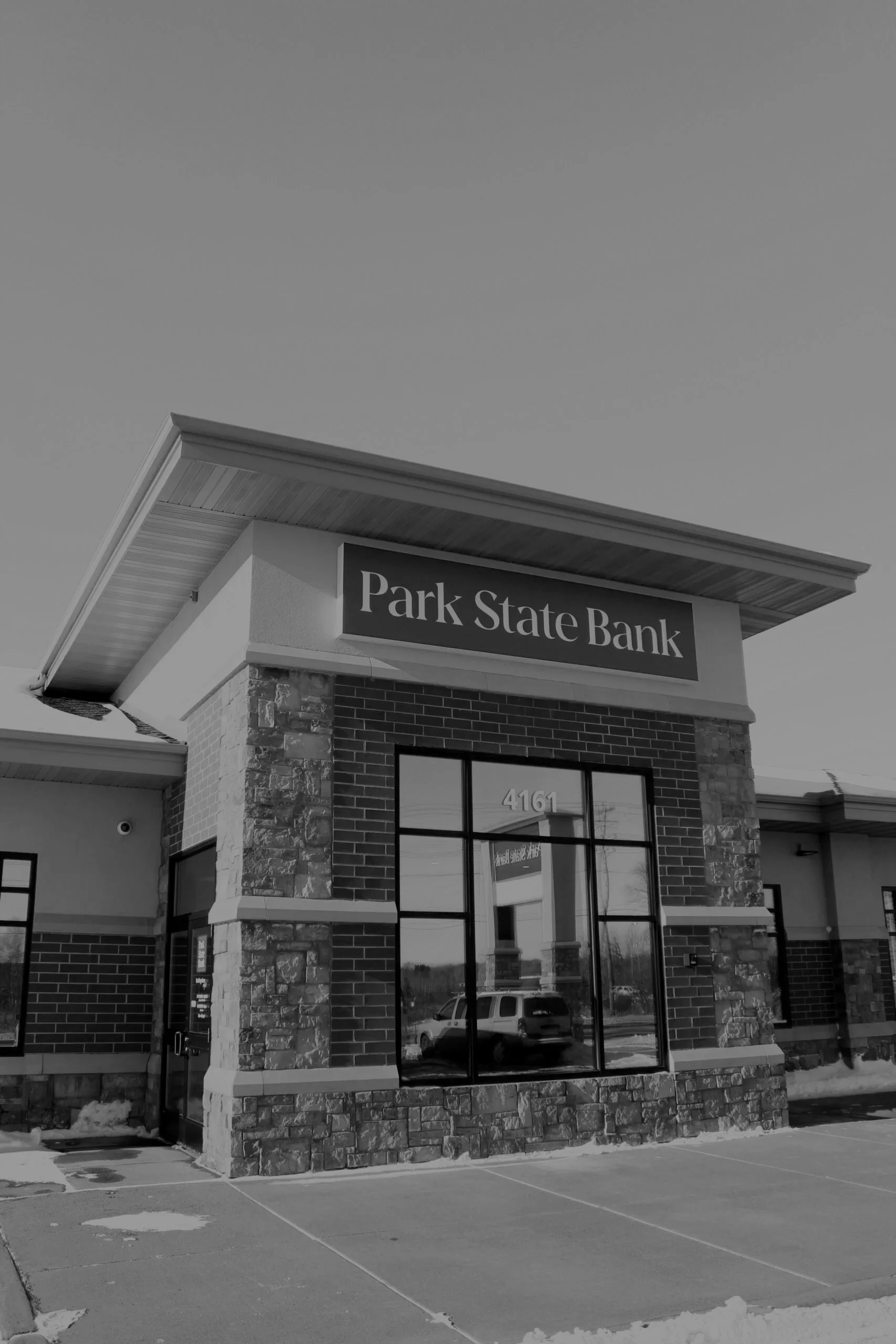 Front view of a modern bank building with a sign that reads 'Park State Bank', large windows, and brick and stone exterior, taken on a sunny day.