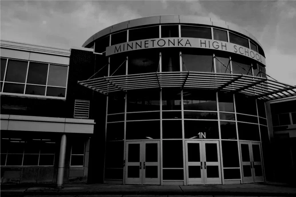 Black and white photo of the front entrance to Minnetonka High School, featuring large glass windows and doors, with an overhanging canopy and a sign displaying the school's name.