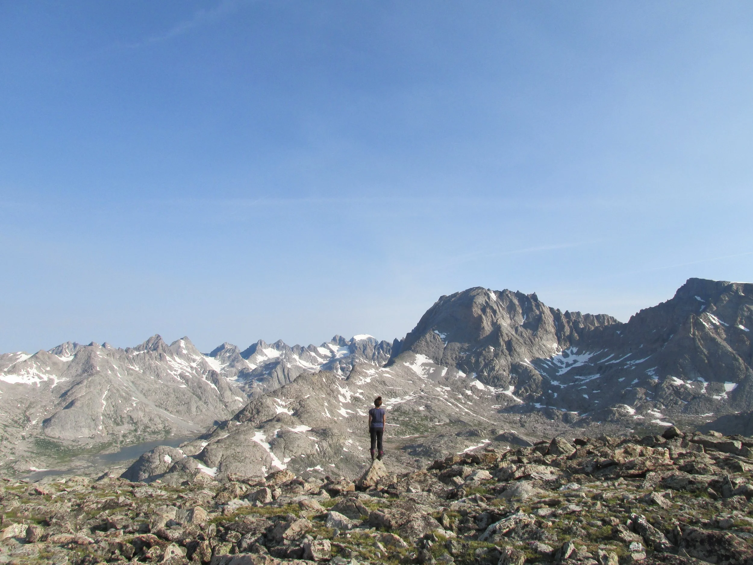 A person standing on rocky terrain in a mountain landscape with snow patches and large mountain peaks under a clear blue sky.