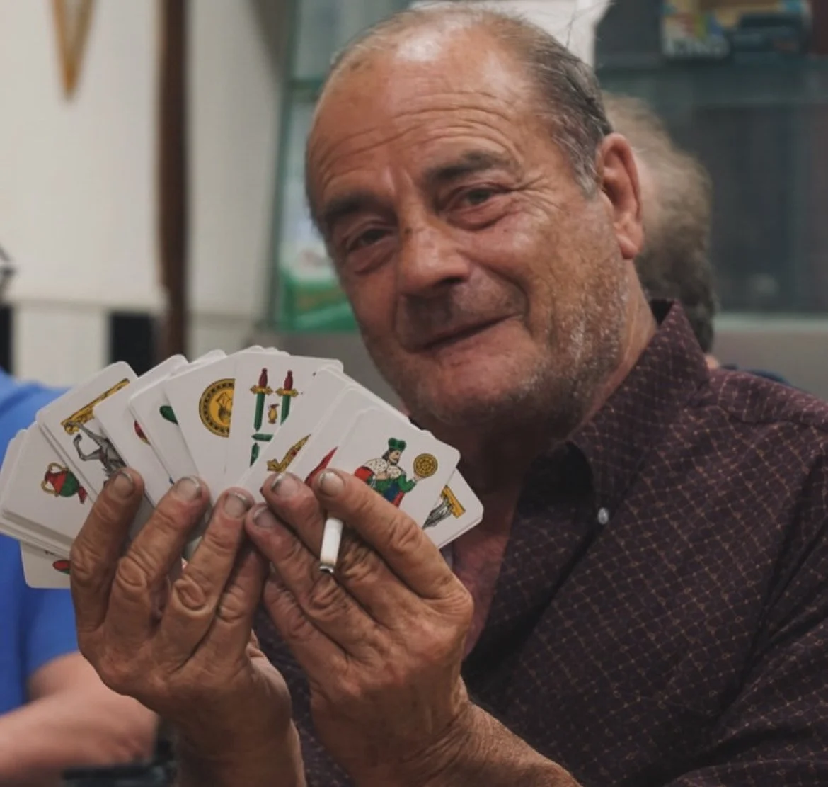 An older man holding a fan of playing cards with various illustrations, smiling slightly in a casual indoor setting.