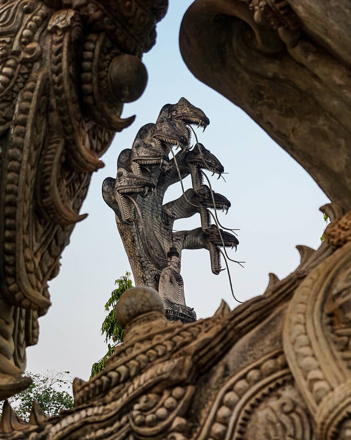A stone sculpture of a multi-headed serpent with an individual standing at the base, viewed through an intricately carved frame, set against a blue sky with some green trees in the background.