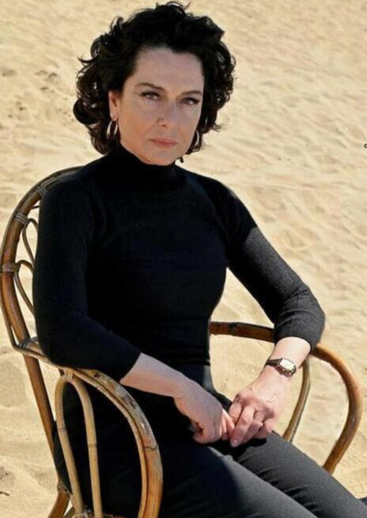 Woman with dark, curly hair wearing a black turtleneck and earrings, sitting on a rattan chair on a sandy beach.