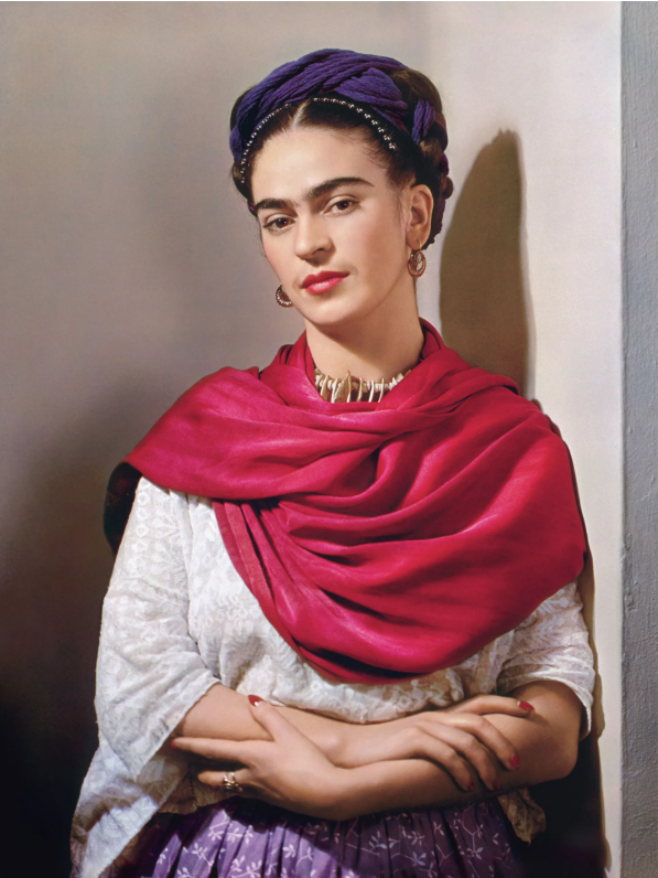 A woman with dark hair styled in a braided headwrap, wearing a purple headband with beads, gold earrings, a metallic necklace, a white embroidered blouse, and a maroon shawl, standing against a plain wall.