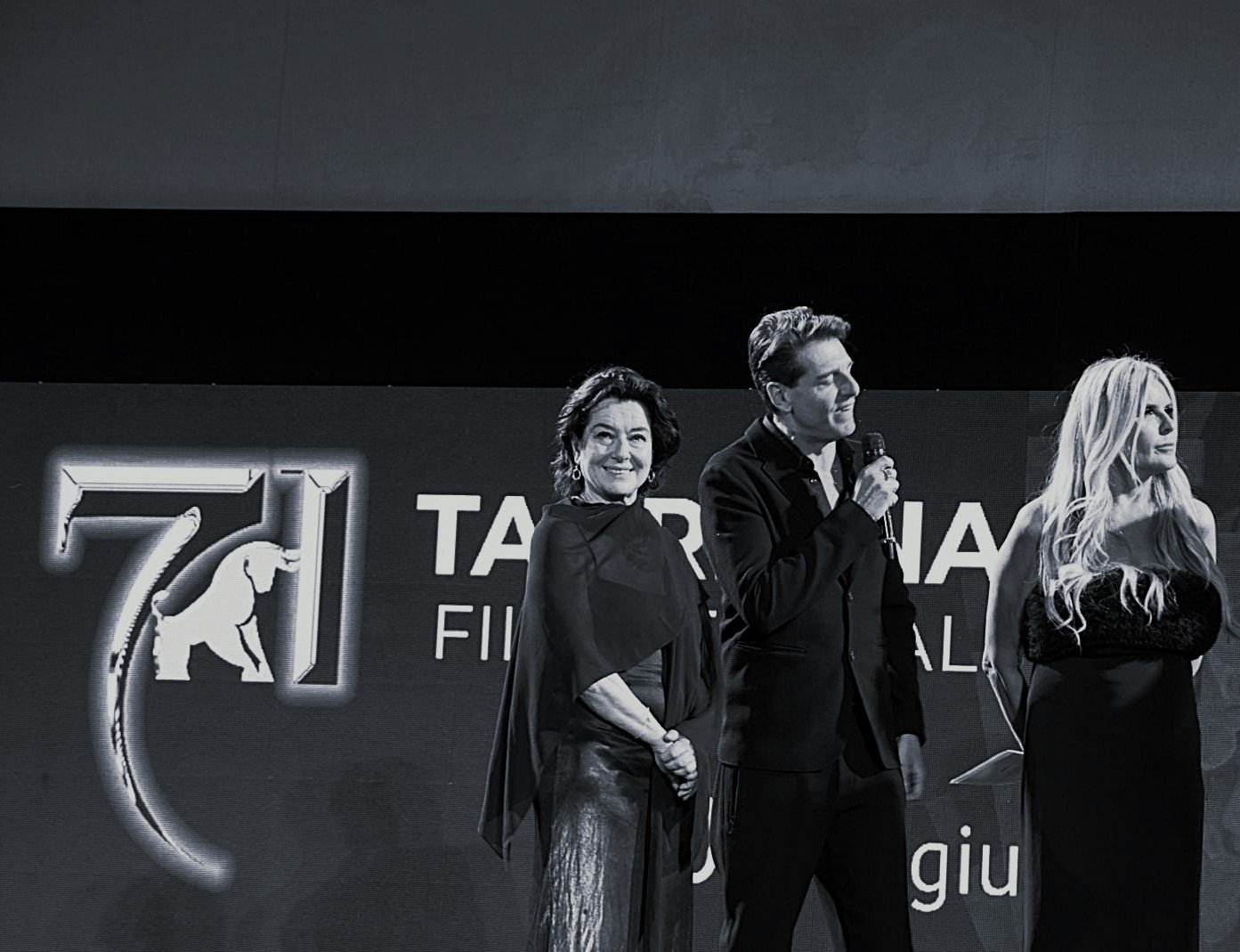 A woman in a black dress holding a microphone and waving, standing next to a man in a suit on stage at an event.