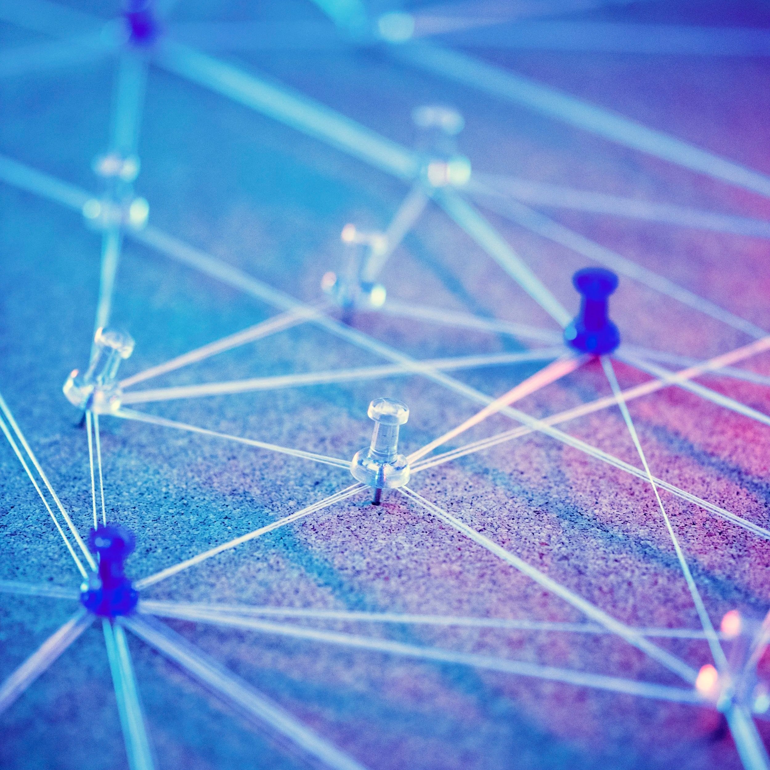Close-up of a network of pins and white threads forming a geometric web on a textured surface, illuminated with blue and pink lighting.