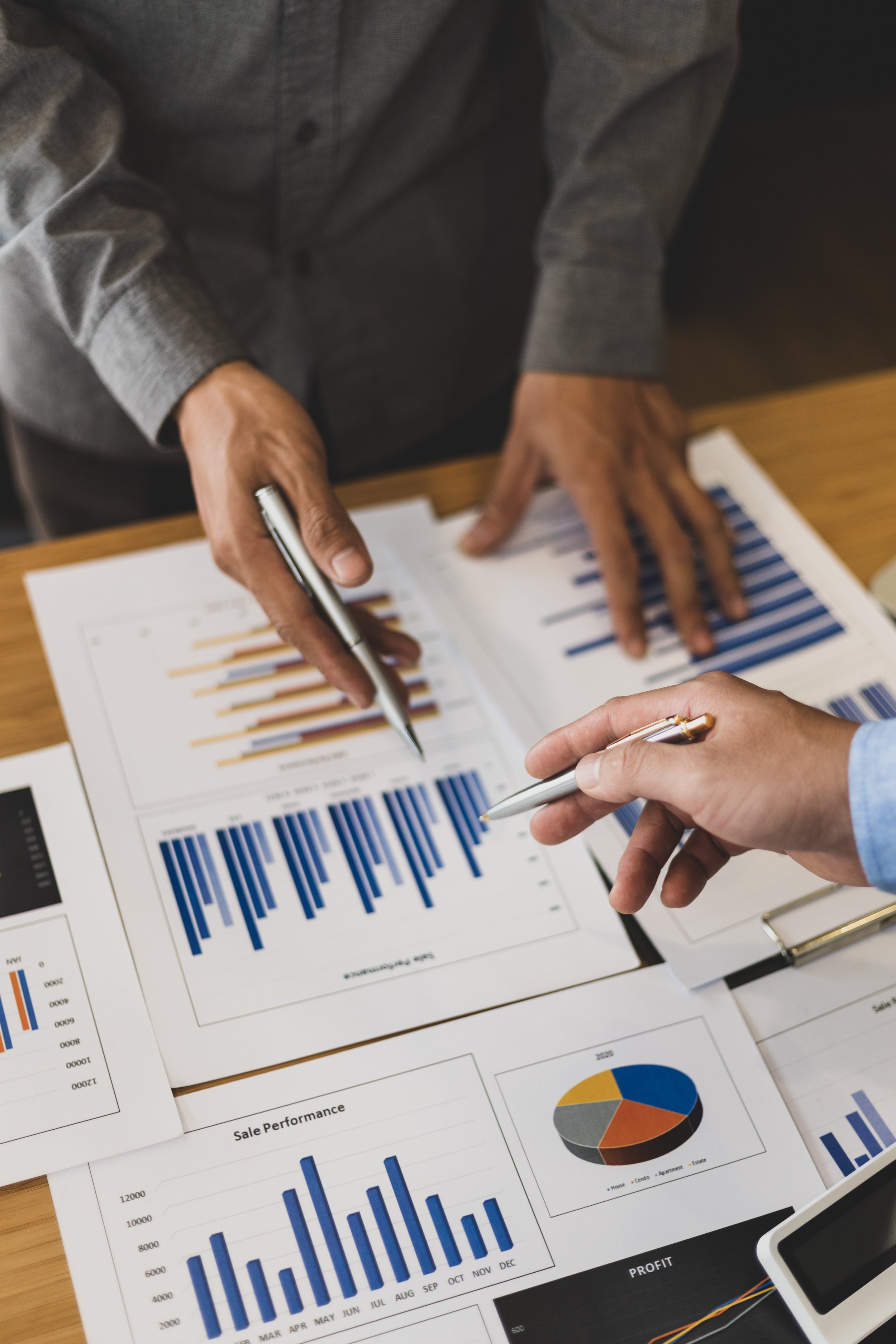 Two individuals analyzing printed sales and financial charts with bar graphs, a pie chart, and data sheets on a wooden table.