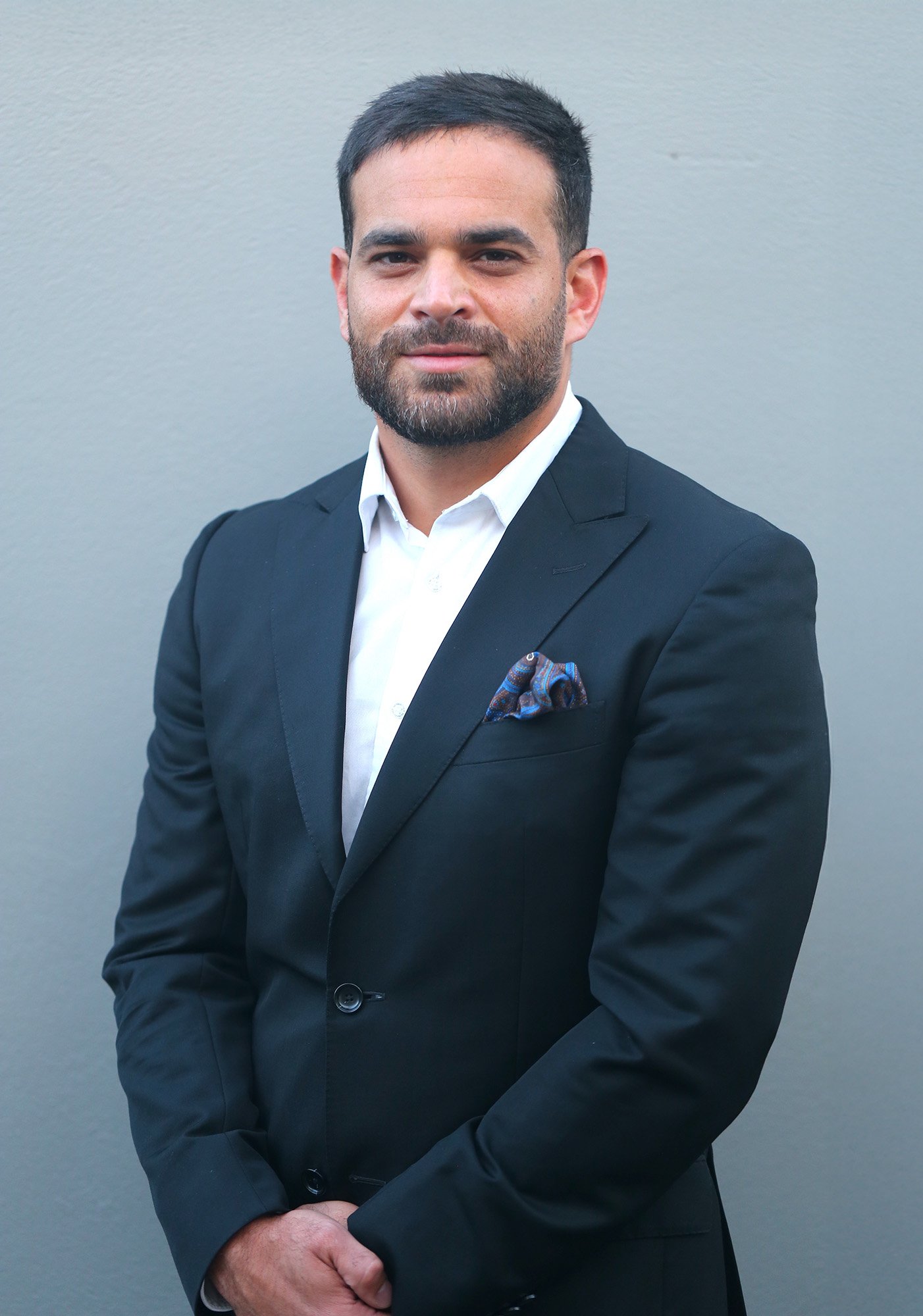 A man with dark hair and a beard dressed in a black suit, white shirt, and pocket square standing against a plain gray background.
