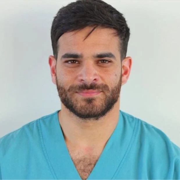 Man with dark hair and beard wearing blue scrubs, smiling against a plain light background.