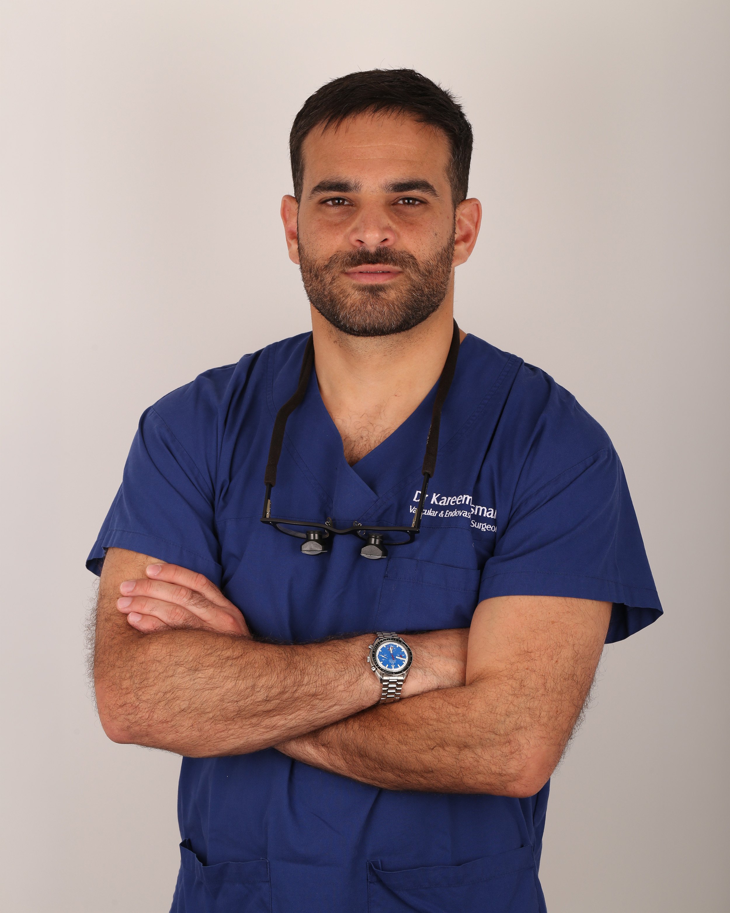 A male medical professional wearing navy scrubs, a wristwatch, and glasses hanging around his neck, standing with arms crossed against a plain white background.