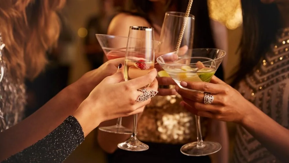 Close-up of people clinking cocktail glasses at a party or gathering, with a background of blurred warm lighting.